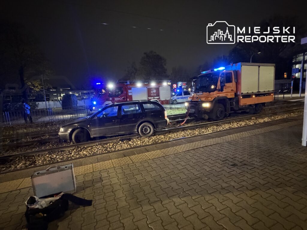 Czarny samochód i pomarańczowa ciężarówka stoją na torowisku, obok służby ratunkowe z włączonymi światłami.