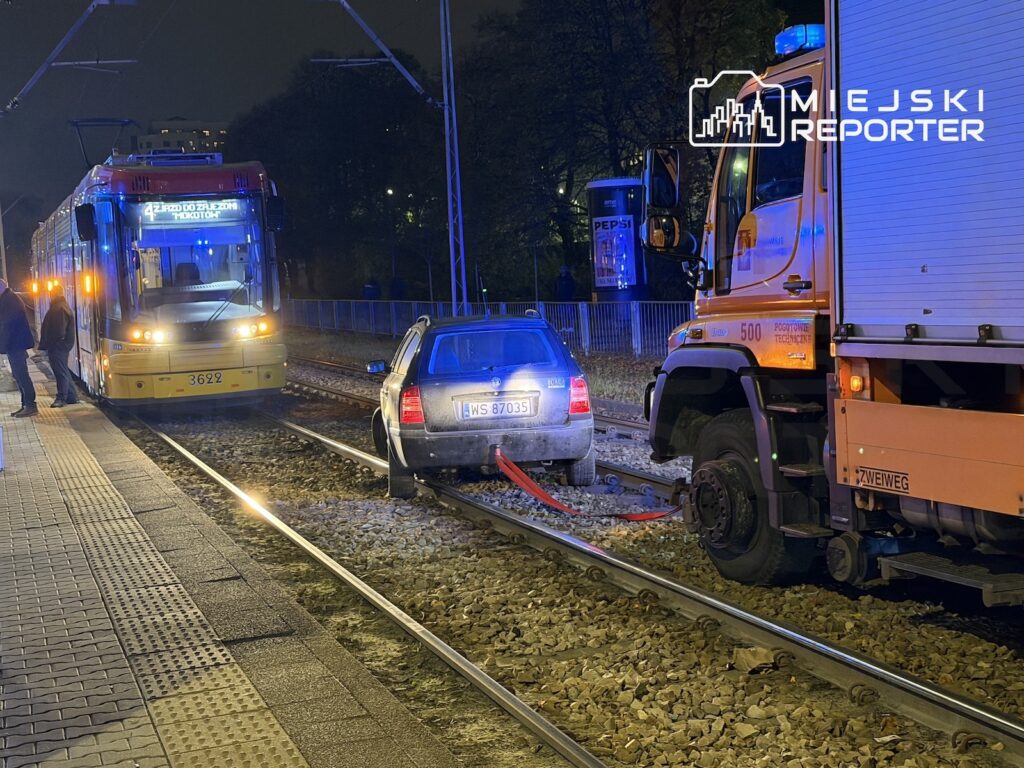 Samochód osobowy stoi na torach tramwajowych między tramwajem a ciężarówką, nocą oświetlony niebieskimi światłami.