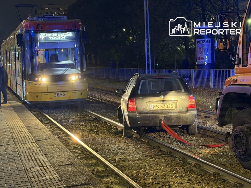 Samochód osobowy stoi na torach tramwajowych przed nadjeżdżającym tramwajem, nocą przy przystanku.