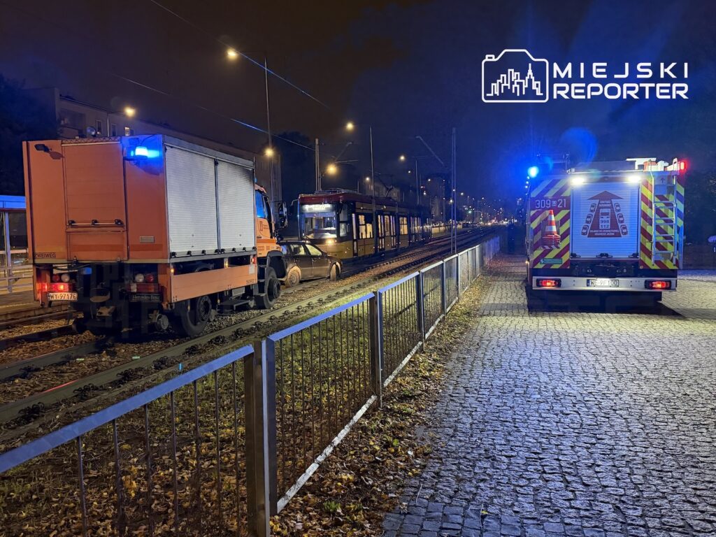 Dwa wozy strażackie z włączonymi niebieskimi światłami stoją nocą przy torowisku, obok tramwaju i samochodu osobowego.
