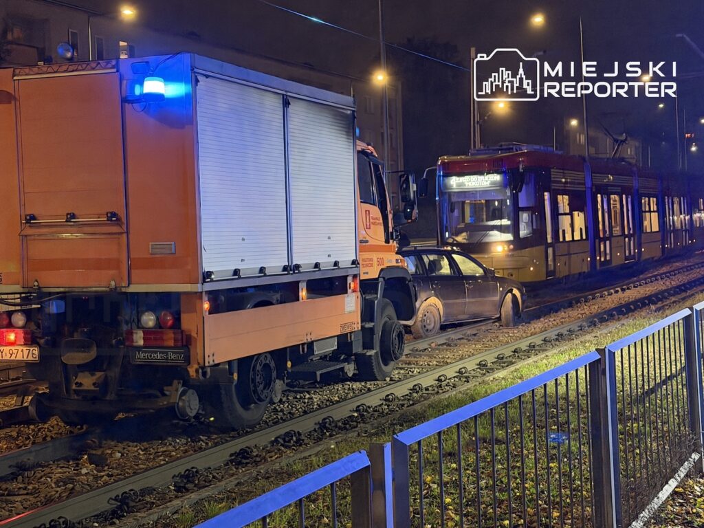 Ciężarówka i samochód osobowy stoją na torowisku przed tramwajem nocą, przy niebieskim świetle sygnalizacyjnym.