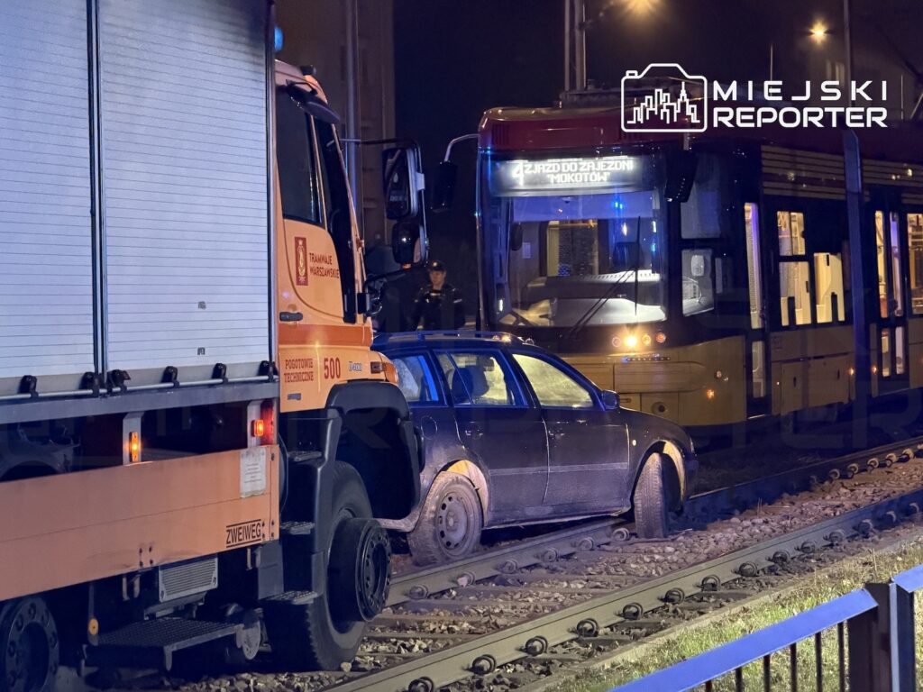Samochód osobowy stoi na torowisku między ciężarówką a tramwajem, nocą przy oświetlonej ulicy.