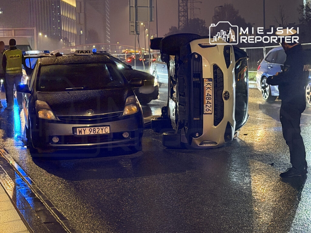 Samochód leży na boku na mokrej jezdni obok taksówki, a policjanci zabezpieczają miejsce zdarzenia nocą.