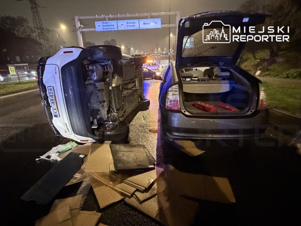 Przewrócony na bok samochód i porozrzucane kartony na mokrej jezdni, obok auta z otwartym bagażnikiem nocą.
