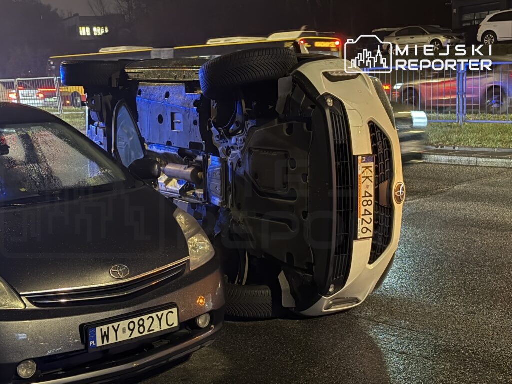 Biały samochód leży na boku przy zaparkowanej Toyocie na mokrej ulicy nocą.