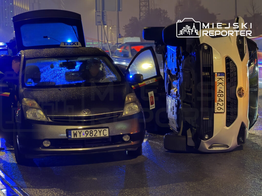 Biały samochód leży na boku obok taksówki z otwartymi drzwiami, na mokrej ulicy nocą.