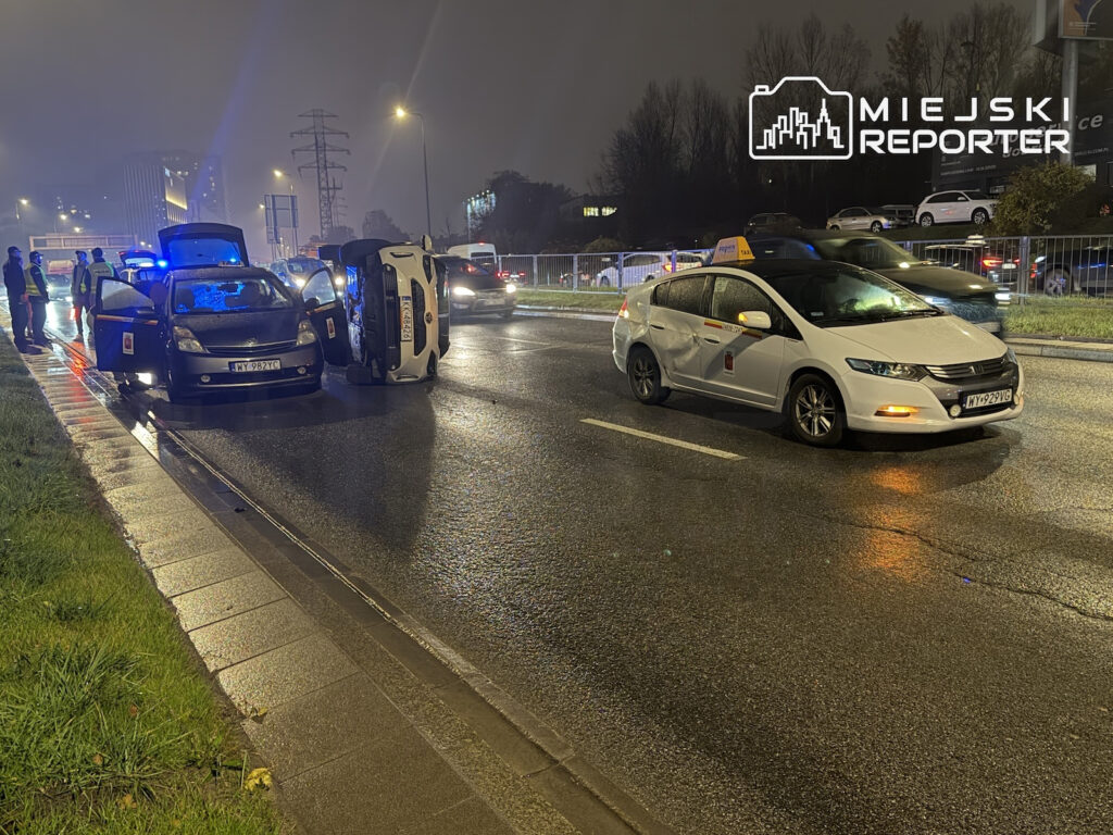 Przewrócony samochód leży na boku na mokrej jezdni, obok stoją auta i pojazd z włączonymi niebieskimi światłami.