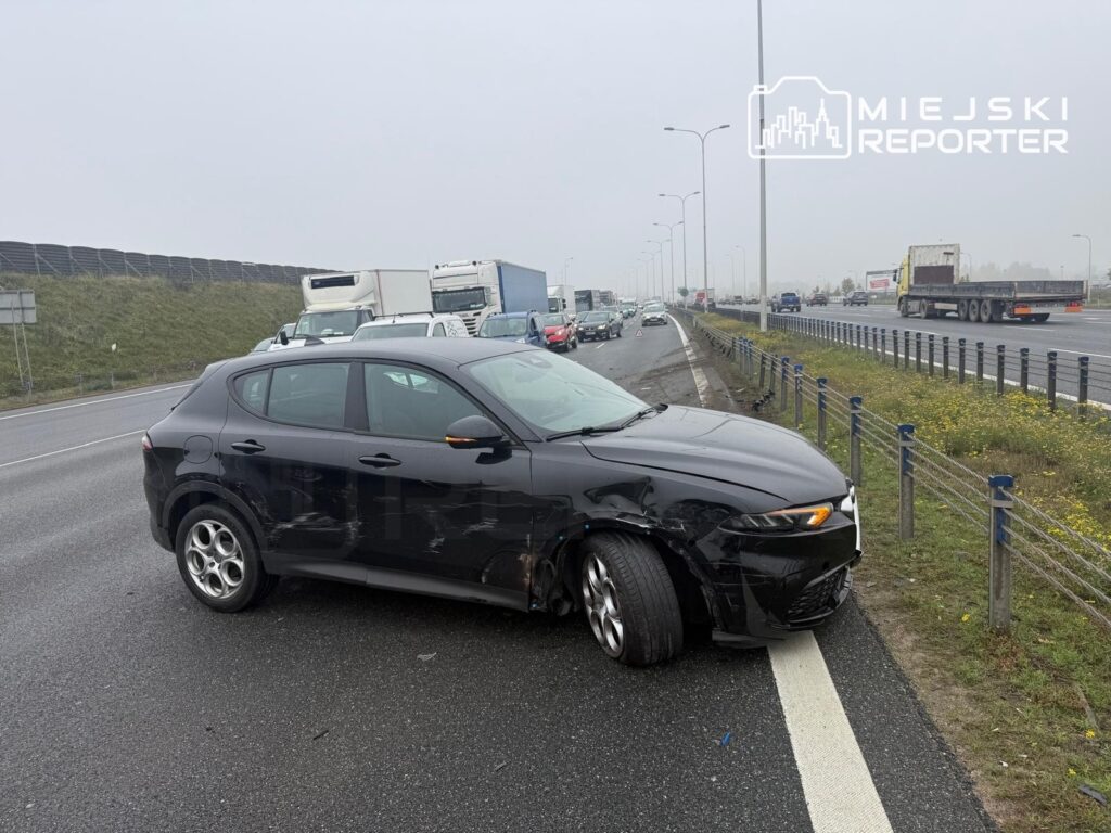 Uszkodzony czarny samochód osobowy stoi ukośnie na poboczu drogi, obok sznura pojazdów i barierek.