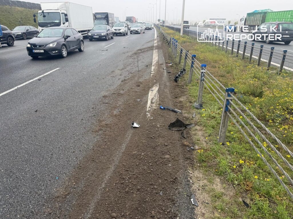 Samochody jadą autostradą obok uszkodzonej bariery i odłamków leżących na poboczu.