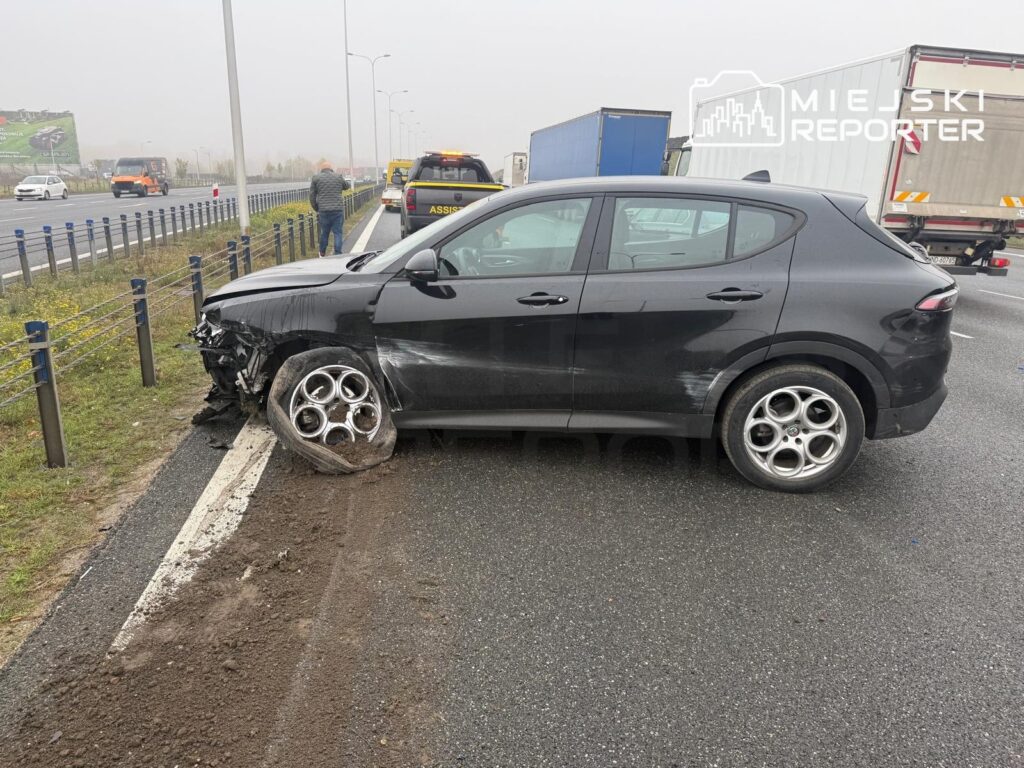 Uszkodzona czarna Alfa Romeo stoi w poprzek pasa ruchu przy barierkach, z rozbitym przodem i uszkodzonym lewym przednim