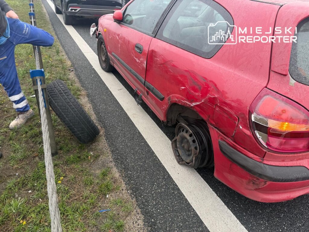 Czerwony samochód z uszkodzonym bokiem i bez tylnego koła stoi na poboczu drogi; obok leży opona.