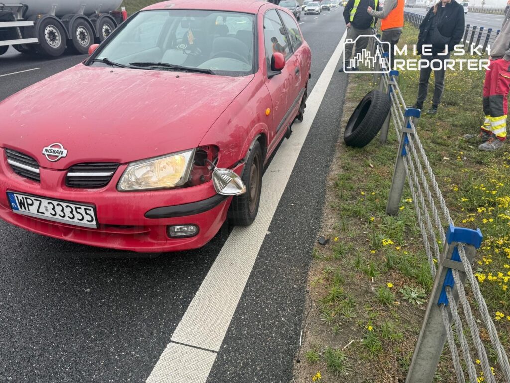 Czerwony Nissan z uszkodzonym przodem stoi na pasie ruchu przy barierkach, obok leży odłączone koło.