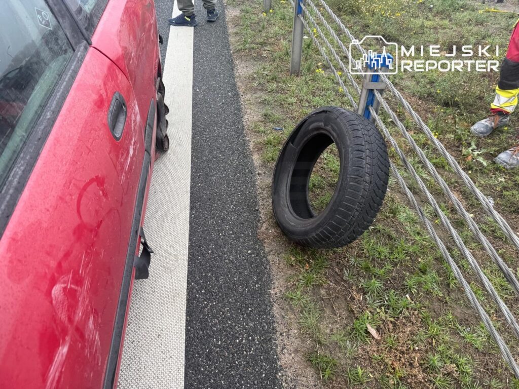 Czerwony samochód stoi przy poboczu, a obok na trawie leży oderwana opona; za nią uszkodzona barierka drogowa.