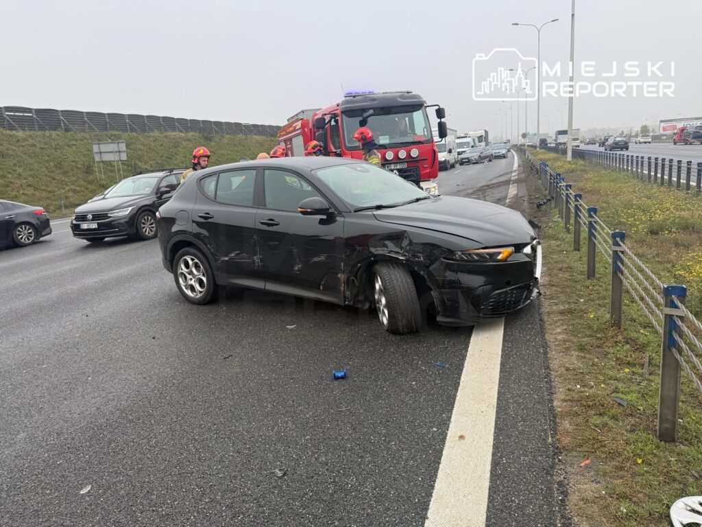 Uszkodzony czarny samochód osobowy stoi na poboczu drogi, obok czerwonego wozu strażackiego i strażaków.