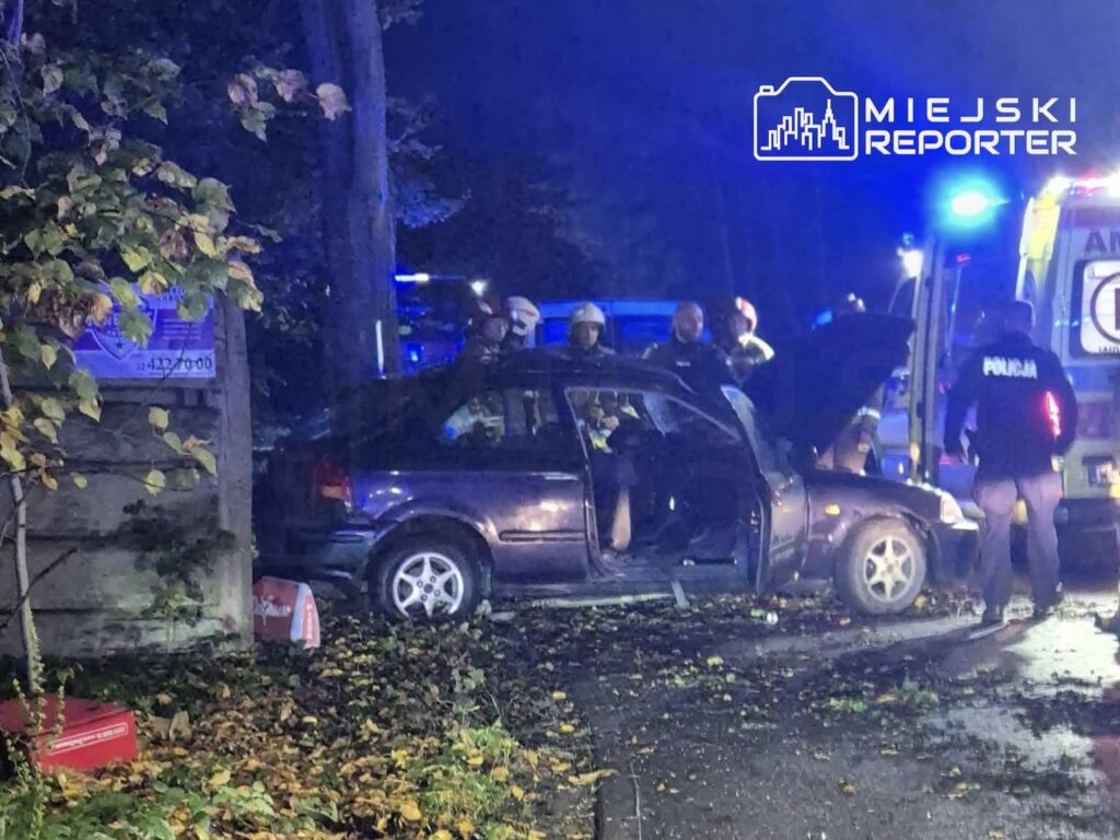 Uszkodzony samochód z otwartymi drzwiami stoi przy drodze nocą, obok służb ratunkowych i policji.
