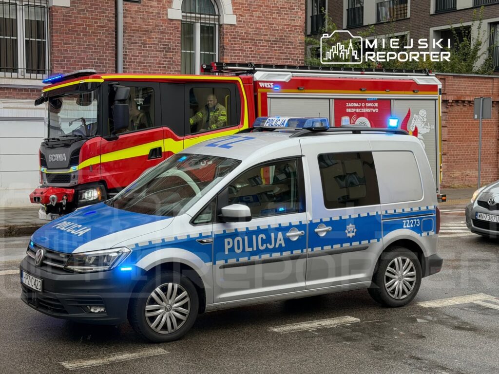 Oznakowany radiowóz policyjny z włączonymi niebieskimi światłami przed czerwonym wozem strażackim na ulicy przy kamienicy.