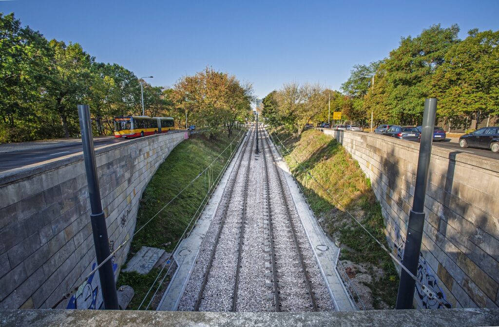 Tory tramwajowe biegną w wykopie między betonowymi murami, obok zieleni i ulicy z samochodami.