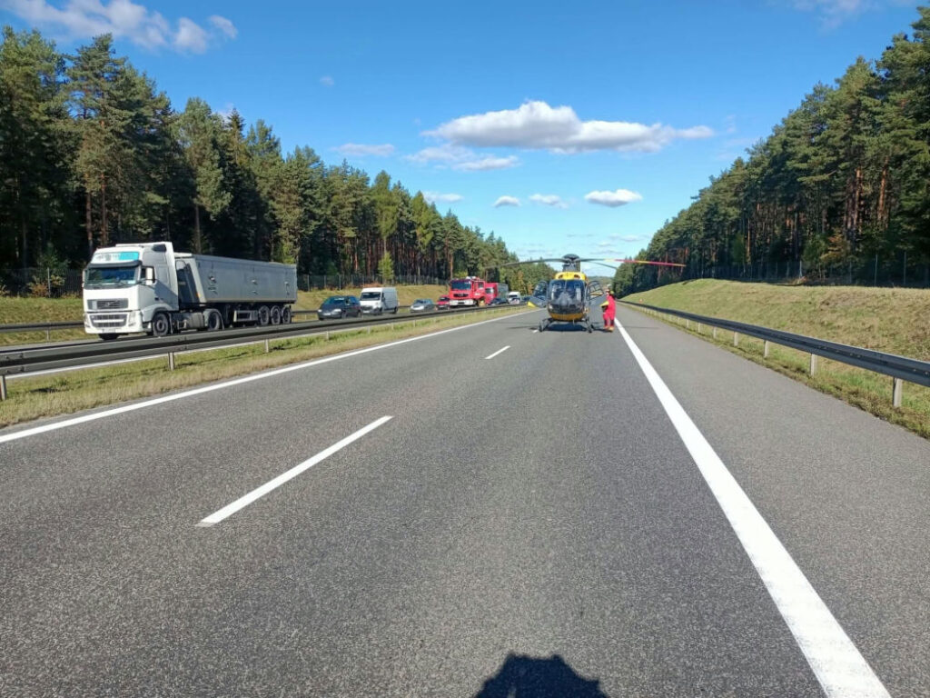 Śmigłowiec ratunkowy i ratownik na pasie autostrady, obok stoją pojazdy służb i tworzy się korek.