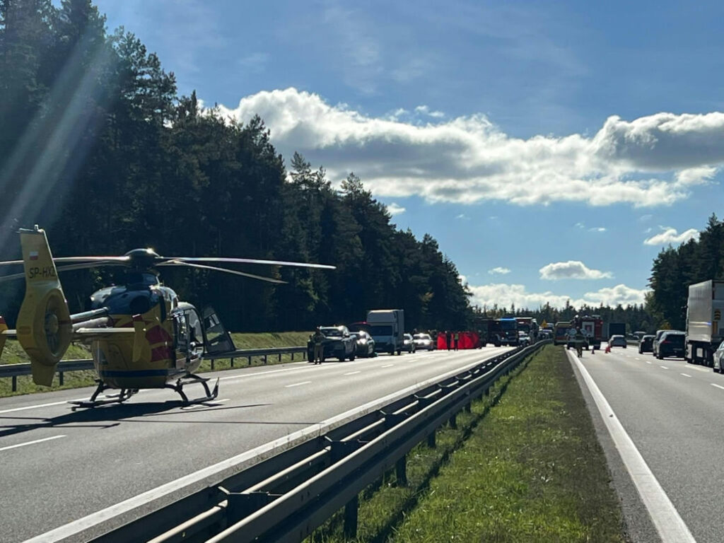 Żółty śmigłowiec stoi na jezdni przy kolumnie samochodów i służb na autostradzie wśród lasów.