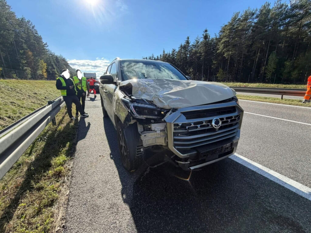 Uszkodzony SUV stoi na poboczu drogi, a obok niego pracują służby w kamizelkach odblaskowych.