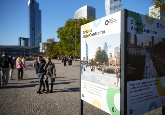 Przechodnie na miejskim placu obok tablicy informacyjnej z napisem „Zielona Aleja Centralna”, w tle wieżowce.