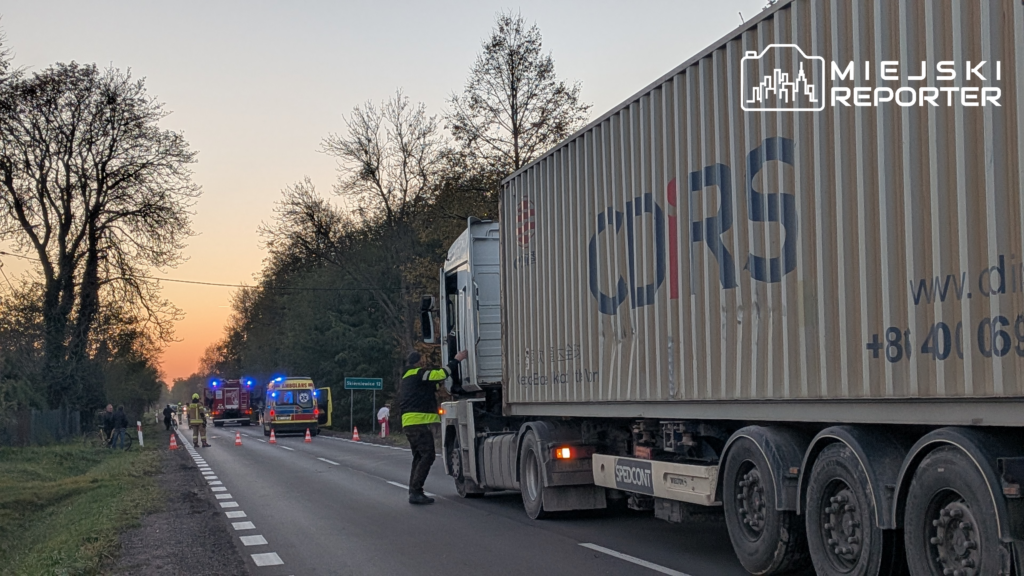 Policjant w kamizelce odblaskowej kontroluje ciężarówkę na drodze, gdzie służby zabezpieczają miejsce zdarzenia.