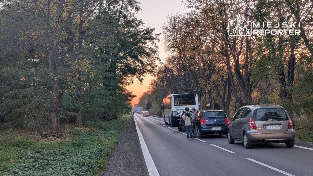 Autobus i samochody stoją na drodze wśród jesiennych drzew, a obok pojazdów znajduje się kilka osób.