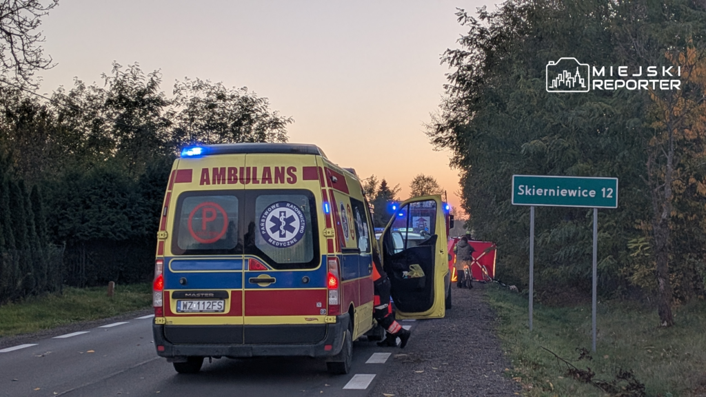 Karetka pogotowia z włączonymi niebieskimi światłami stoi na poboczu przy znaku „Skierniewice 12”.