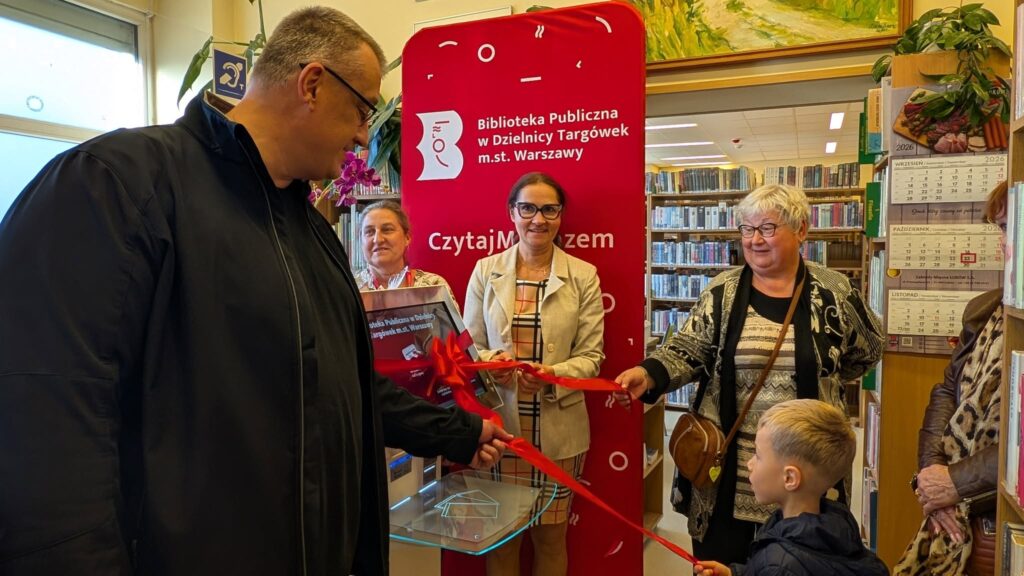 Mężczyzna i kobieta przecinają czerwoną wstęgę w bibliotece, w obecności pracownic i dziecka.