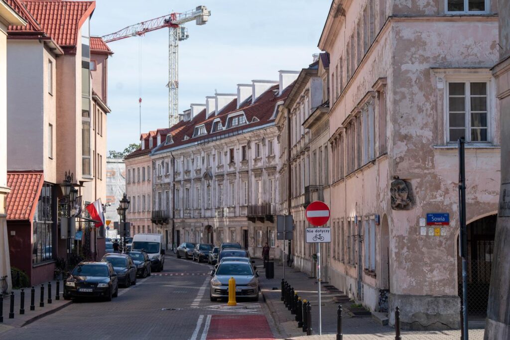 Wąska ulica z zabytkowymi kamienicami, zaparkowanymi samochodami i znakiem zakazu ruchu przy chodniku.