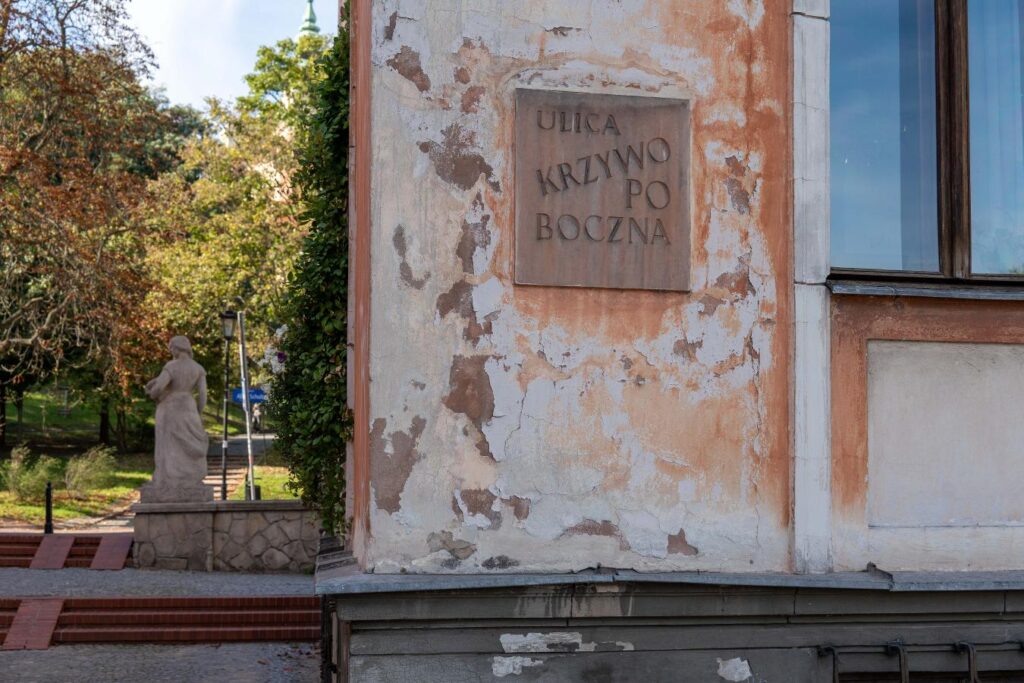 Zniszczona elewacja budynku z tablicą „ULICA KRZYWOPODBoczna” obok parku z kamiennym pomnikiem.