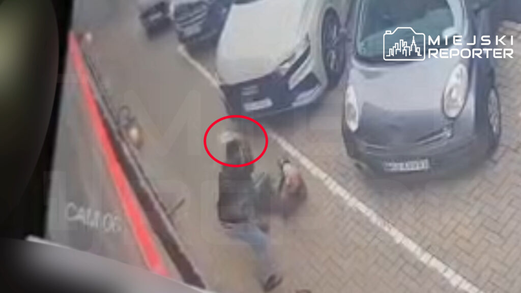 Rozmazane nagranie z monitoringu: dwie osoby na chodniku między zaparkowanymi samochodami, jedna leży na ziemi.