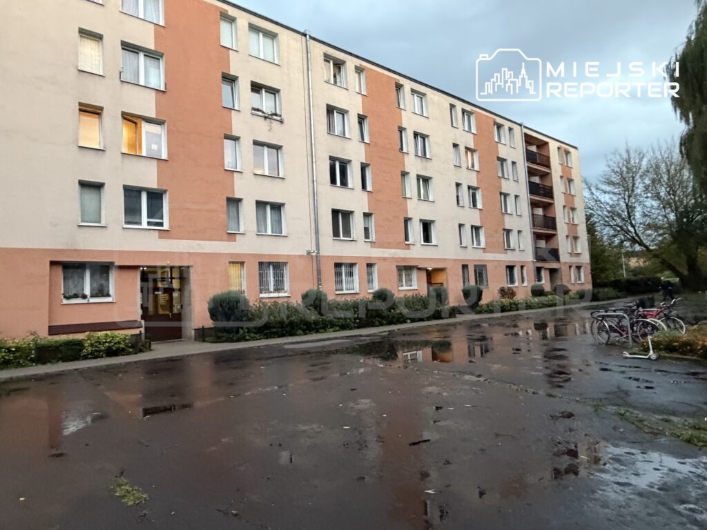 Pięciokondygnacyjny blok mieszkalny przy mokrym, brukowanym placu, obok którego stoją rowery.