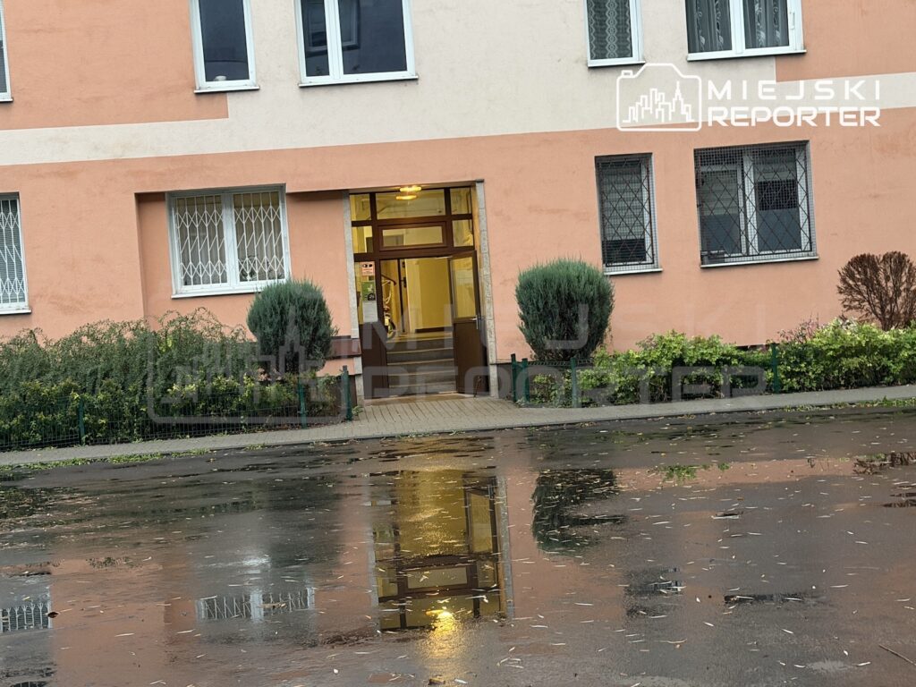 Wejście do różowo-beżowego budynku z okratowanymi oknami odbija się w kałuży na mokrym chodniku.