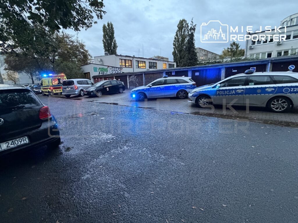 Radiowozy policji z włączonymi niebieskimi światłami i karetka stoją na mokrej ulicy przy budynkach.