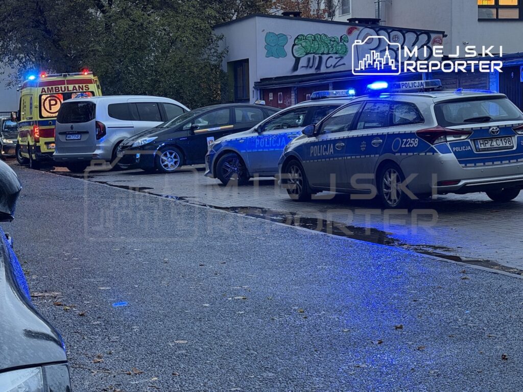Radiowóz z włączonymi sygnałami świetlnymi i karetka stoją na mokrej ulicy przy budynku.