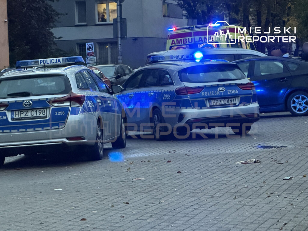 Dwa radiowozy z włączonymi niebieskimi światłami i ambulans stoją nocą na parkingu przy budynku mieszkalnym.