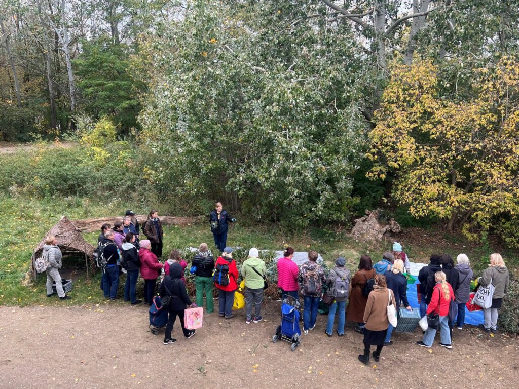 Grupa ludzi słucha przewodnika w parku, otoczona drzewami i krzewami.